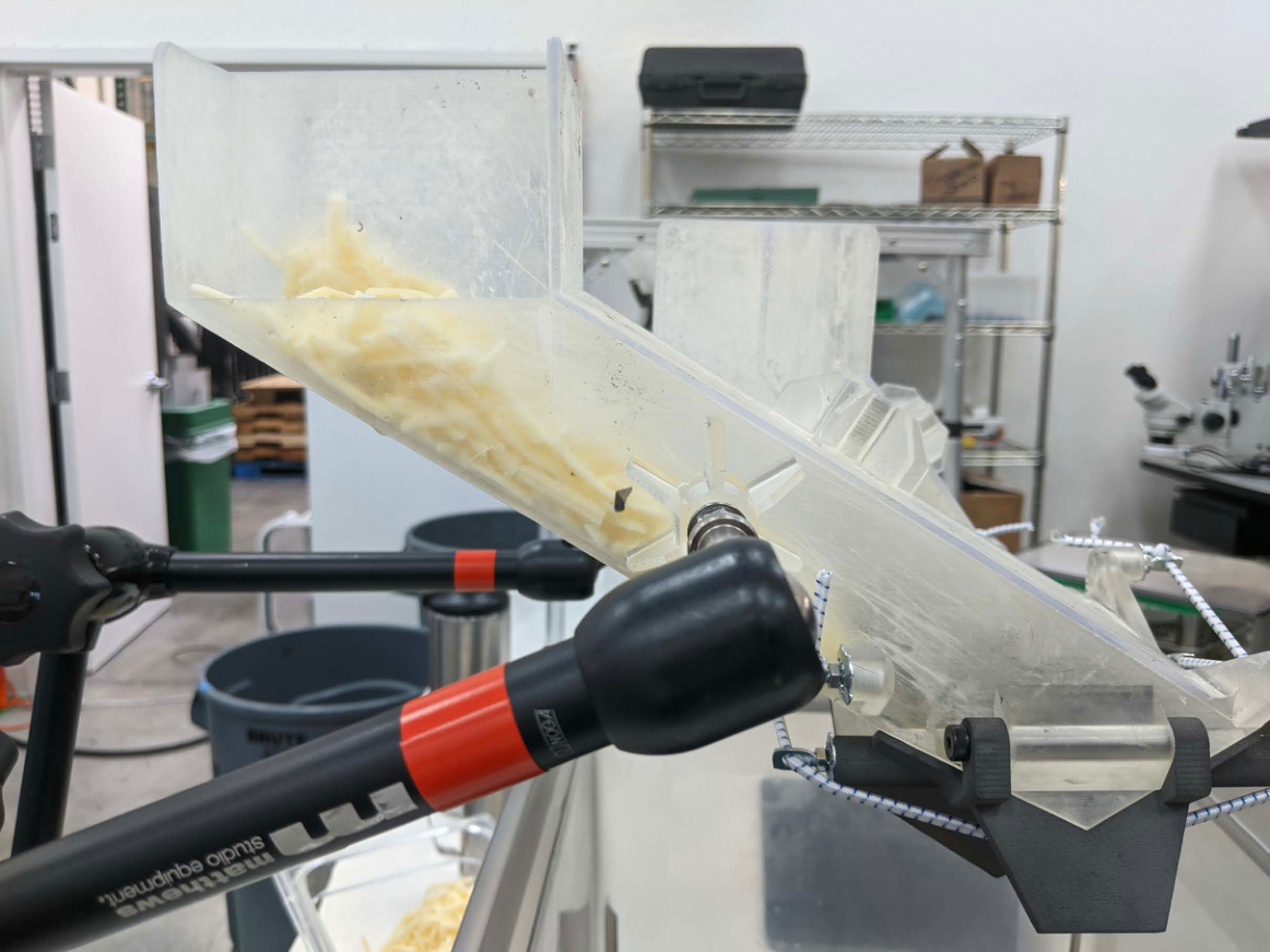 Side view of a transparent clamped chute holding shredded cheese over a collection pan.