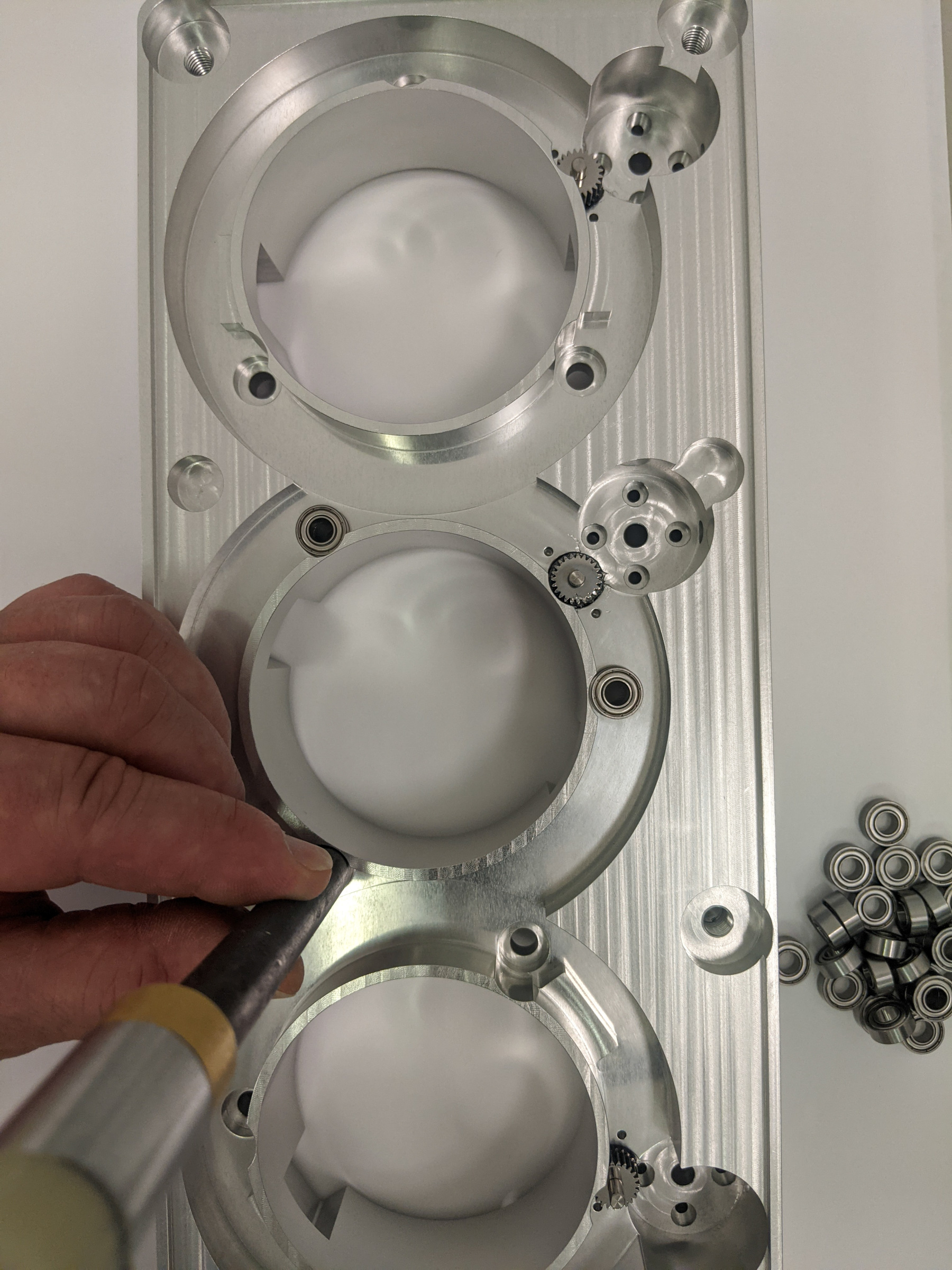 A hand tool works beside bearings and pinions in a machined enclosure with multiple circular cup openings.